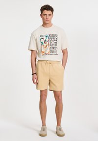 Kerman oversized t-paita, jossa on värikäs graafinen printti, yhdistettynä vaaleankeltaisiin tekstuuriin shortsit. Vaaleat lenkkarit täydentävät asun.