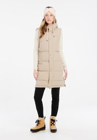 Gilet matelassé beige avec une capuche, des boutons sur le devant, deux poches latérales, porté par-dessus une chemise à manches longues claire et associé avec des leggings noirs et des bottes beiges.