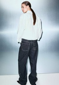 Jeans de mezclilla oscura de corte holgado con adornos metálicos, combinados con una camisa blanca de gran tamaño que presenta un diseño minimalista.