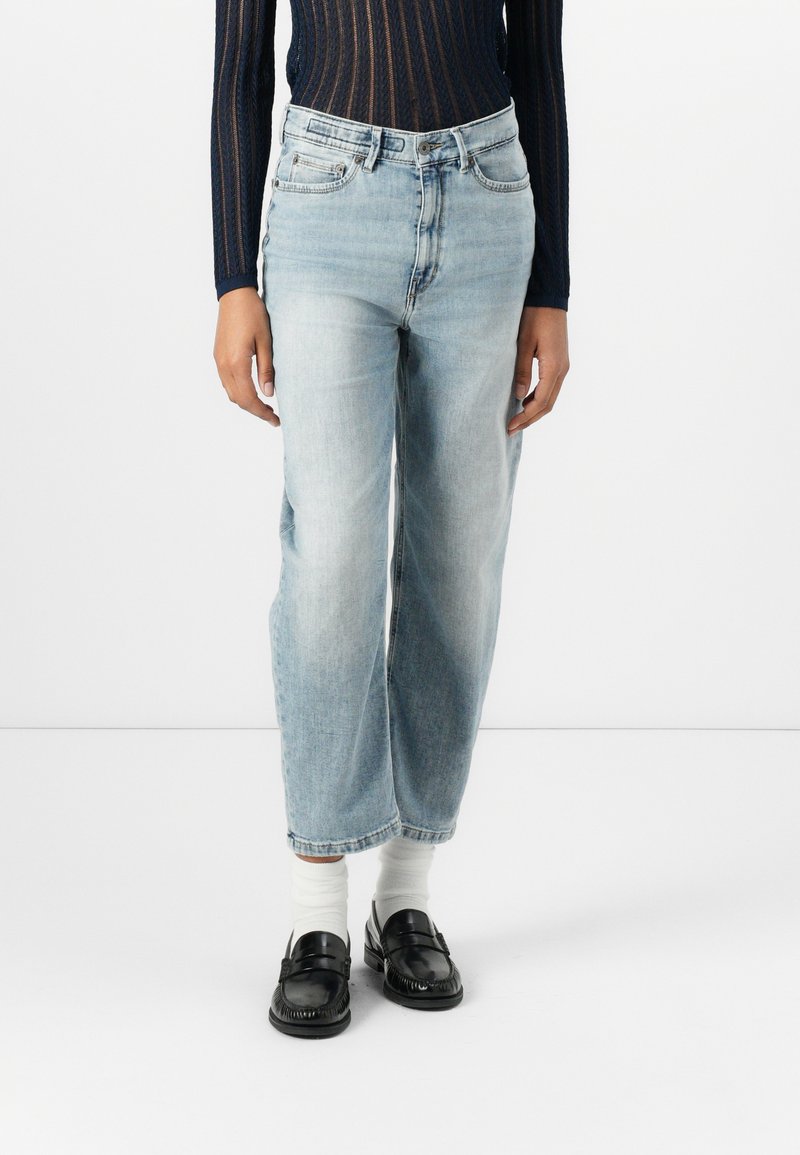 Jeans in denim leggermente blu con una vestibilità rilassata, dotati di una vita alta, tasche frontali e una lunghezza leggermente accorciata. Indossati con mocassini neri.