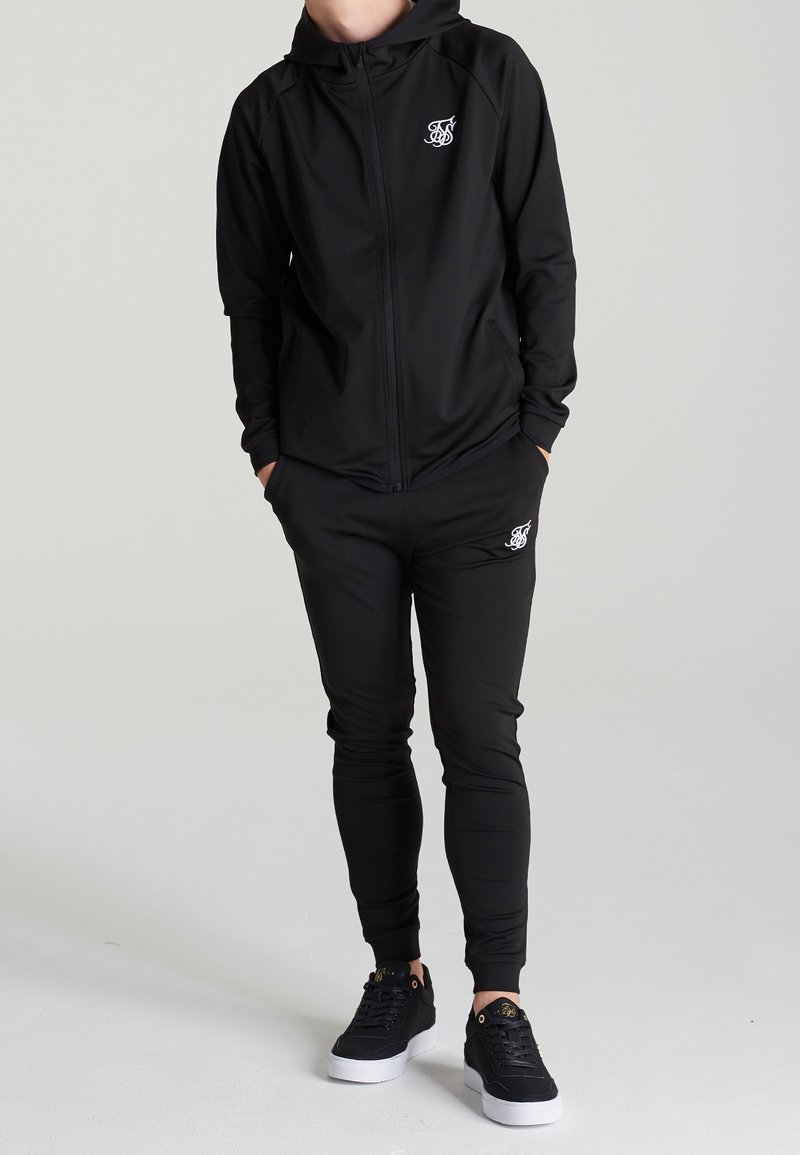 Survêtement noir avec sweat à capuche zippé et pantalon fuselé, présentant un logo blanc sur la poitrine et la cuisse. Tissu lisse et extensible avec une coupe ajustée.
