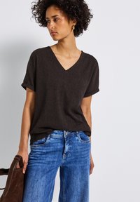 Lösa, kortärmade V-hals topp i mörkbrunt texturerat tyg, tillsammans med ljusblå jeans. Modellen håller en brun väska.