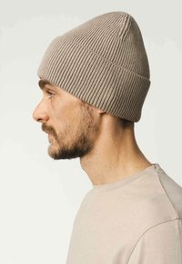 Ribbelige beige gebreide beanie met een taps toelopende top, gedragen op het hoofd van een man, gecombineerd met een lichtgekleurde t-shirt. Zachte textuur, strakke pasvorm.