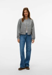 Chaqueta gris corta con cuello redondo y cierres de botón, combinada con jeans de campana azules y botas negras hasta el tobillo. Diseño simple y texturizado.