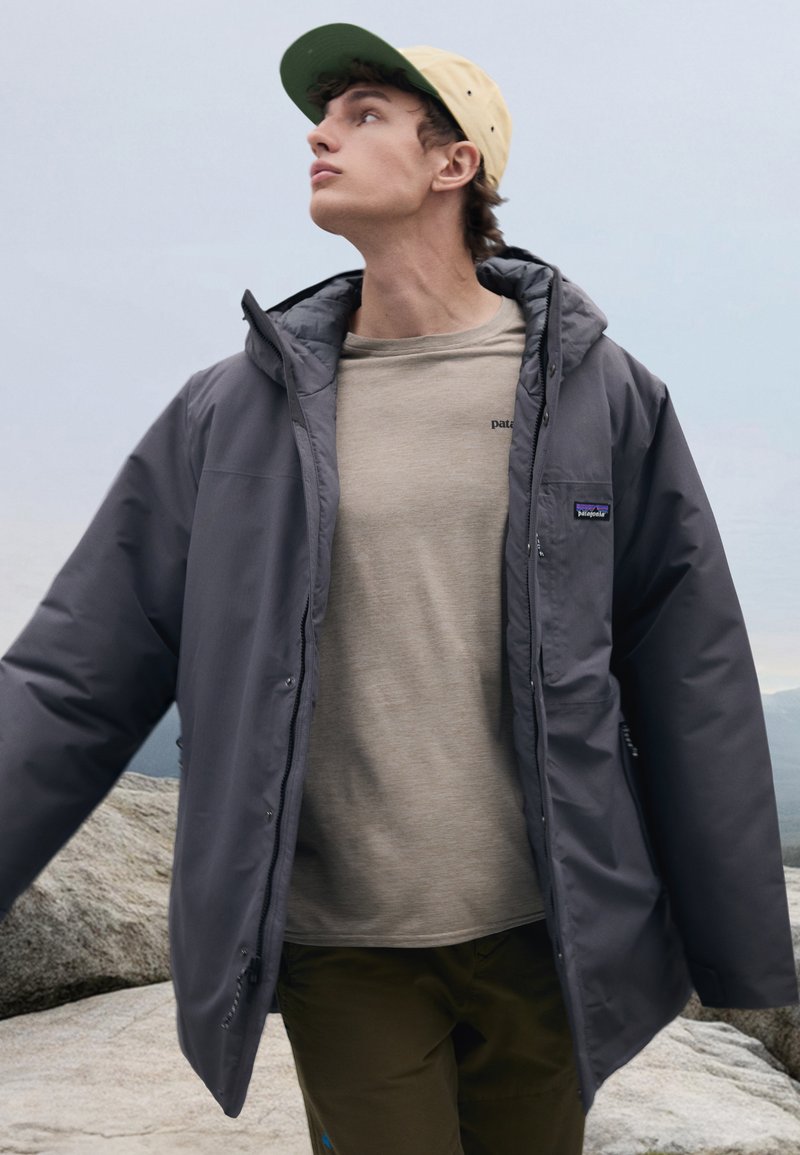 Veste isolée grise avec capuche, dotée d'une fermeture éclair à l'avant et de poches latérales, portée sur une chemise beige clair et un pantalon vert olive.