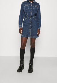 Denimkleid mit langen Ärmeln, Knopfleiste, zwei Brusttaschen und tailliertem Schnitt. Getragen mit knielangen schwarzen Socken und kräftigen schwarzen Stiefeln.