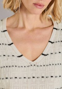 Frau mit kurzem blonden Haar, die lange Kristallohrringe trägt und einen weißen Strickpullover mit schwarzen horizontalen Streifen und einem V-Ausschnitt.