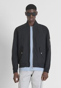 Giubbotto bomber nero con collo rialzato, zip frontale e due tasche laterali. Presenta una tasca con zip sulla manica e una texture morbida.