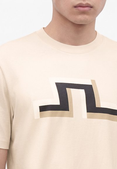 Μπεζ T-shirt με μαύρο, άσπρο και μπεζ γεωμετρικό ζικ-ζακ σχέδιο στο στήθος, φορεμένο από ένα άτομο που φαίνεται από τον λαιμό έως τον άνω κορμό.