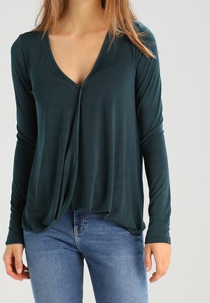 Maglietta lunga azzurra con scollo a V e davanti drappeggiato. Realizzata in un tessuto morbido con una vestibilità rilassata, abbinata a jeans blu.