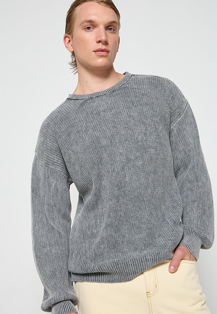 Jongeman in een grijze gebreide trui en lichtbeige broek, staand met een hand in zijn zak tegen een effen achtergrond.