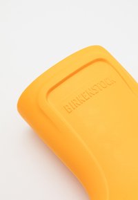 Sole in gomma arancione con superficie texturizzata, caratterizzata da branding in rilievo "BIRKENSTOCK" e un bordo liscio, angolata per la prospettiva.