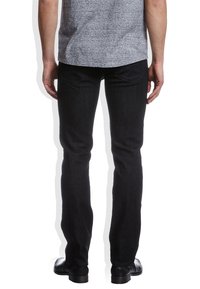 Jeans negros ajustados con una textura suave y detalles de costura sutiles, con bolsillos traseros, usados con una camisa gris y zapatos negros.