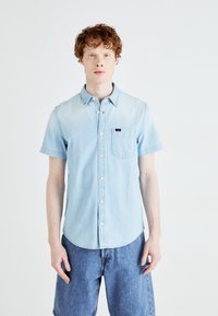 Chemise en denim bleu clair à manches courtes et à boutons, avec une poche poitrine et une étiquette logo discrète. Associée à un short en denim bleu.