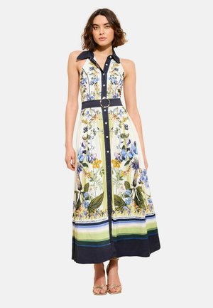 Kobieta ubrana w bezrękawową, zapinaną na guziki sukienkę maxi z nadrukiem kwiatów i motyli, z granatowym kołnierzem, paskiem i paskowanym dołem, w połączeniu ze złotymi sandałami.