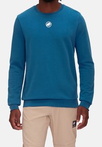 Blå bomullssweatshirt med rund halsringning, långa ärmar och en cirkulär vit logotyp på bröstet. Slät textur, åtsittande mudd och fåll.