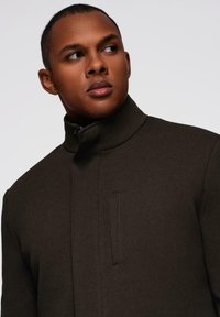 Chaqueta de color marrón oscuro con cuello alto y tejido texturizado, que presenta un cierre de cremallera y un detalle de bolsillo lateral. Diseño moderno y estructurado.
