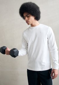 Langærmet hvid skjorte med rund hals, kombineret med sorte bukser. Figuren holder sorte sekskantede håndvægte, sat mod en lys baggrund.