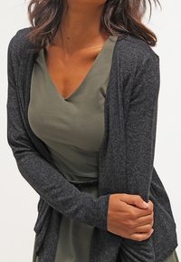 Žena v olivově zelených šatech s výstřihem do V a tmavě šedém kardiganu s otevřeným předním dílem, která drží jednu ruku druhou.