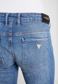 Acercamiento al bolsillo trasero de unos jeans de denim azul claro con el logotipo metálico triangular de Guess y la etiqueta de cuero negro de Guess en la cintura.