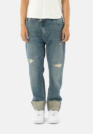 Lichtblauwe versleten denim jeans met een losse pasvorm, voorzien van rafelige hems en grote patches. Gecombineerd met witte sneakers.