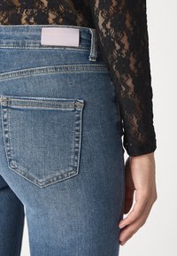 Jeans de denim azul ajustados con un lavado medio, que cuentan con un único bolsillo trasero y una etiqueta blanca en la cinturilla. Parte superior de encaje negro texturizado visible.