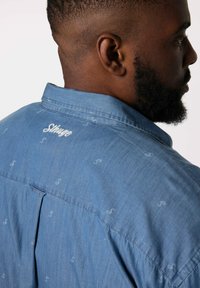 Blå denimskjorta med subtila mönster av dollartecken, spetsig krage och broderat varumärke på baksidan. Mjuk känsla, avslappnad passform.