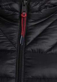 Zbliżenie na czarną pikowaną kurtkę z centralnym zamkiem błyskawicznym, z czarnym uchwytem zamka oraz przywieszką z plecionki w kolorze czerwonym i granatowym.