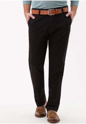 Pantaloni di cotone neri con design a gamba dritta, abbinati a una cintura di pelle marrone. Le scarpe sono marroni con lacci. Lo sfondo è bianco semplice.