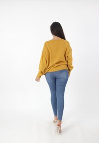 Mosterdgele oversized gebreide trui met verlaagde schouders en geribbelde manchetten, gecombineerd met aansluitende blauwe spijkerbroeken. Heldere achtergrond.