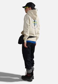 Felpa beige con cappuccio e grafica sul retro, pantaloni cargo neri, calzini grigi e stivali neri robusti con dettagli rossi. Berretto nero e occhiali da sole.