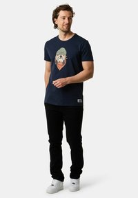 Marinblå bomullströja med en grafik av en hund i en grön mössa. Kombinerad med svarta jeans och vita sneakers, stående mot en neutral bakgrund.