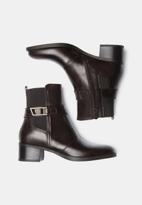 Bottines en cuir marron avec une fermeture éclair sur le côté, un panneau élastique, un talon bloc et des détails en métal sur la sangle. Texture lisse avec une finition brillante.