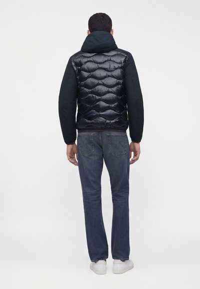 Homme debout de dos portant une veste matelassée noire avec une capuche, un jean bleu et des baskets blanches sur un fond blanc uni.