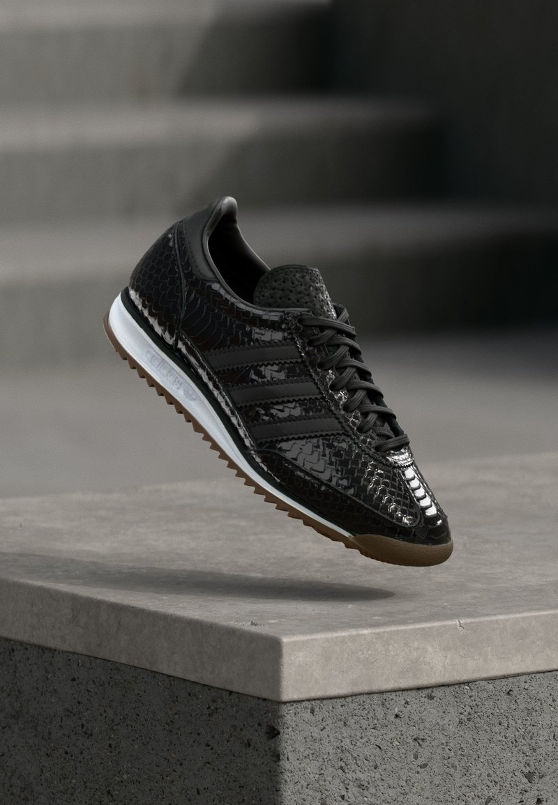 Baskets noires brillantes avec motif texturé, lacets noirs, semelle intermédiaire blanche et semelle extérieure marron, présentées au-dessus d'un rebord en béton.