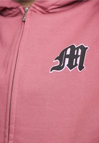 Sudadera con cremallera de color rosa y textura suave. Presenta un logo gráfico en blanco y negro en el pecho. Cremallera con acabado metálico.