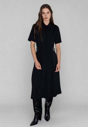Donna che indossa un vestito midi nero a maniche corte con dettagli fibbia laterali e stivali neri alti al ginocchio con punta affilata, in piedi davanti a uno sfondo grigio.