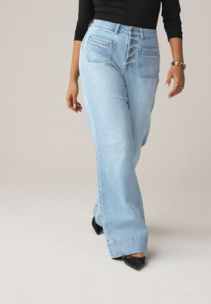 Femme portant un jean large taille haute bleu clair avec des boutons à l'avant, des chaussures noires pointues, un top noir et des bijoux au poignet droit et aux doigts.