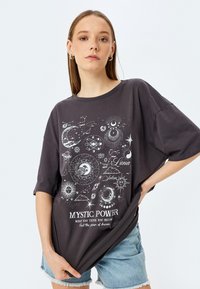 T-shirt oversize gris foncé en coton, avec un imprimé graphique céleste en blanc, comprenant des lunes, des étoiles et le texte "POUVOIR MYSTIQUE."