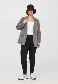 Blazer grigio con rever a notched, due tasche frontali, abbinato a un top bianco, leggings neri e scarpe da ginnastica bianche. Cappellino nero indossato.