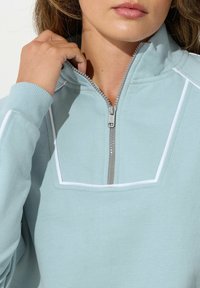 Pullover azzurro chiaro con colletto a zip a quarto, bordo bianco a contrasto e polsini a coste. Tessuto morbido con una texture liscia.