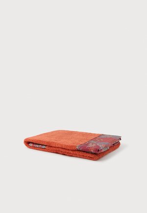 Serviette orange pliée avec un bord rouge et bleu à motifs reposant sur une surface grise claire unie.