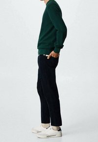 Pull côtelé vert, pantalon sombre et baskets blanches. Le pull présente un col rond et des manches longues ; le pantalon est ajusté avec une poche arrière.