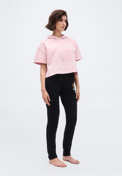 Γυναίκα με κοντά καστανά μαλλιά, φοράει cropped ροζ φούτερ με κουκούλα και μαύρο παντελόνι με μικρό σχέδιο αρκουδάκι, στέκεται ξυπόλυτη πάνω σε απλό φόντο.