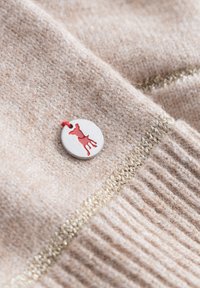 Beige brei stof met een geribbelde textuur, voorzien van een ronde label met een ontwerp van een rode jurk en een rode draad; goudkleurig accent stiksel zichtbaar.