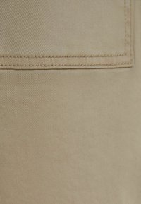Béžová látka s hladkým povrchem, obsahující obdélníkovou náprsní kapsu a viditelné švy podél okrajů. Jednoduchý design.