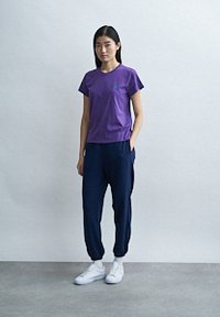 Μωβ κοντομάνικο μπλουζάκι με στρογγυλή λαιμόκοψη, συνδυασμένο με ναυτικό μπλε παντελόνι φόρμας με πλαϊνές τσέπες και ελαστικές μπατζάκια, που φοριέται με λευκά αθλητικά παπούτσια.