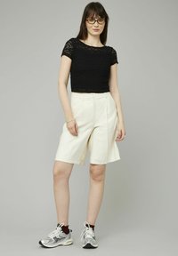 Top court en dentelle noire, shorts ivoire haute jusqu'aux genoux, et chaussures de sport argentées. Le mannequin se tient avec une main sur la hanche, le fond est gris neutre.