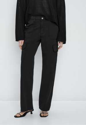 Persoon draagt zwarte wijd uitlopende cargobroek met klepzakken aan de voorkant, een loszittende zwarte gebreide trui en zwarte sandalen met hak en bandjes.