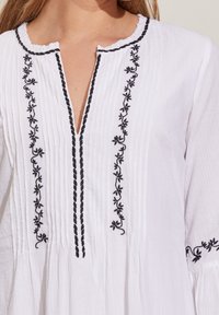 White blouse with a textured fabric, featuring black floral embroidery along the neckline and central placket, and gathered sleeve accents.